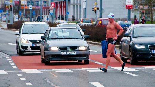 Muž bez trička, jen v plavkách a koupací čepici, přechází po přechodu pro chodce