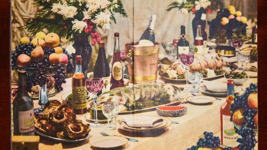 Snímek z klasického vydání sovětské kuchařky z roku 1955, na němž je vyobrazena bohatá, přepychová večeře se šampaňským, pečeným prasetem a dalšími pokrmy