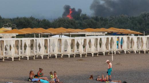 Lidé odpočívají na pláži, zatímco po explozích na ruské vojenské letecké základně v Novofedorivce na Krymu stoupá kouř a jsou vidět plameny, 9. srpna 2022