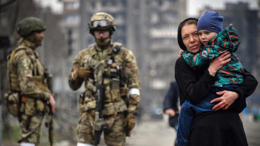 Žena drží své dítě v ulicích Mariupolu, opodál postávají ruští vojáci